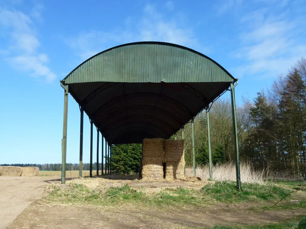 An open-sided Dutch barn with a curved roof and a stack of bales under it