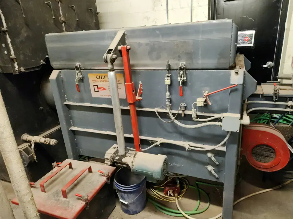 A grey wood-chip boiler with its feed and controls in a school boiler room