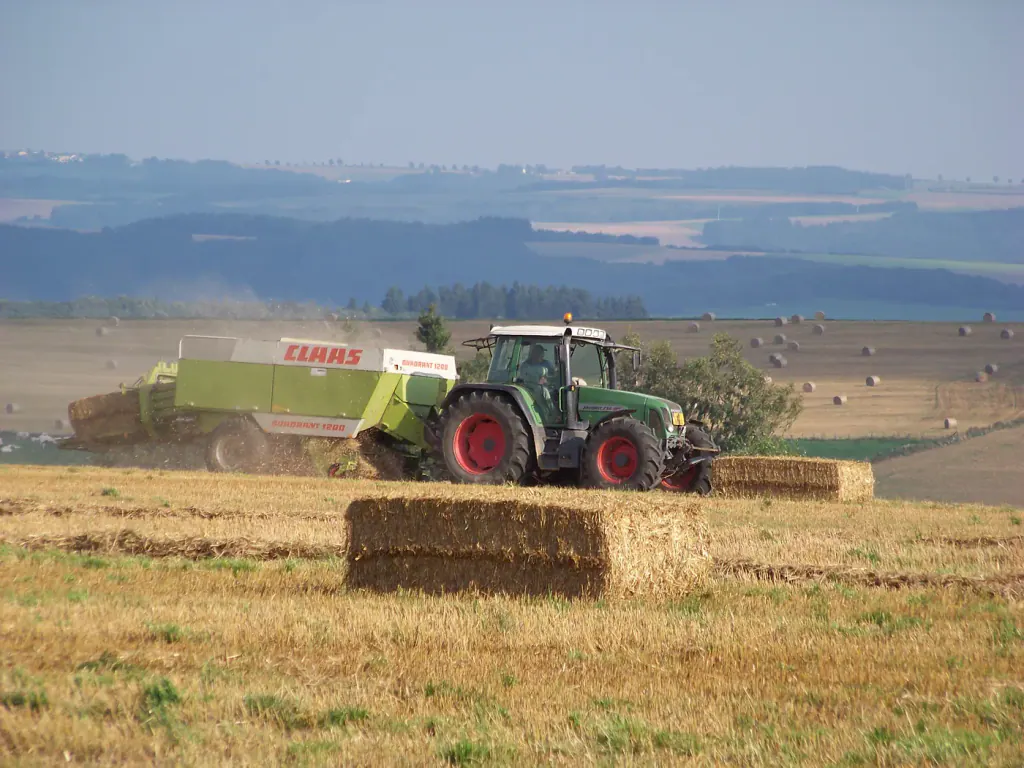 A green tractor pulling a big square baler across a straw stubble field, with a finished bale in front
