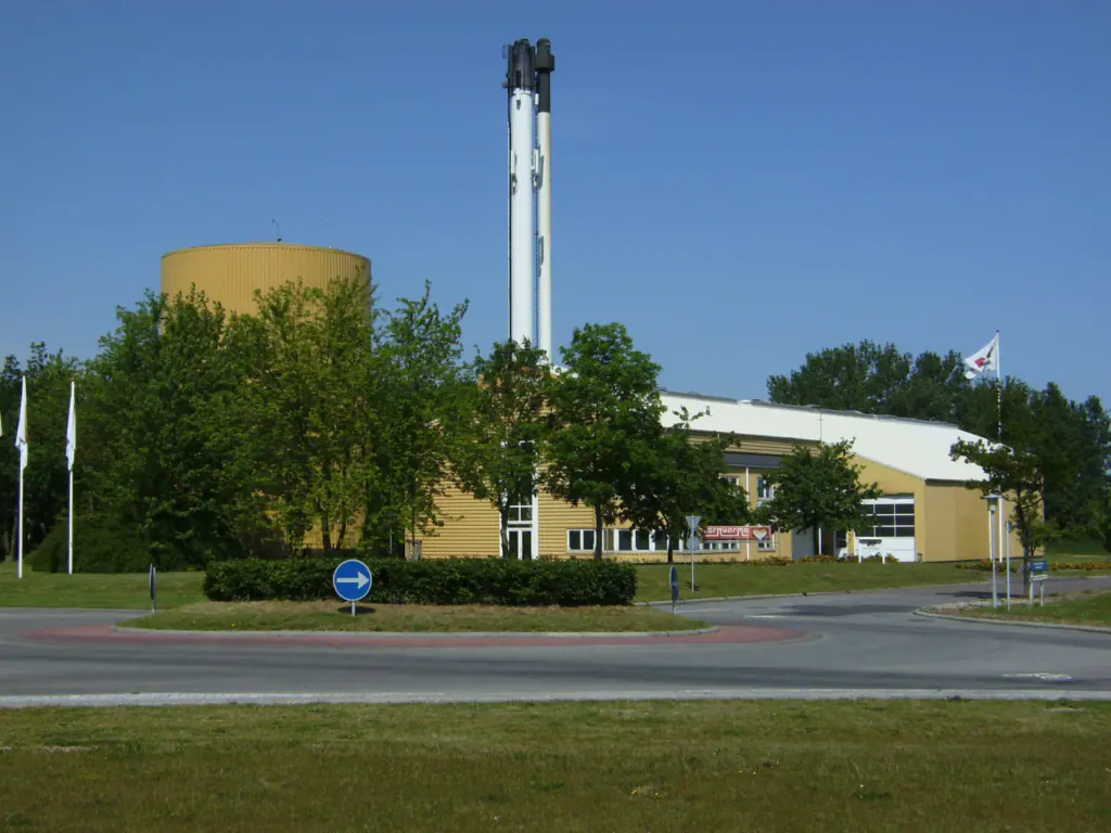 A straw-fired district heating plant with a tall chimney and a round storage tank behind a roundabout