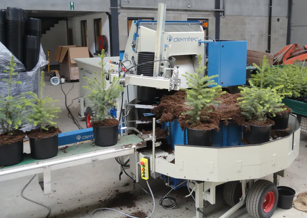 A potting machine indoors filling black containers with growing medium around young conifers
