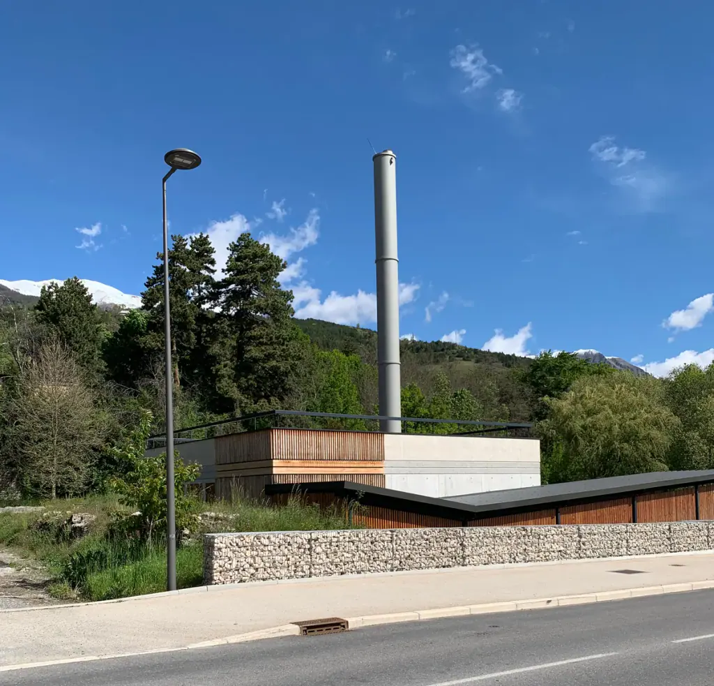 A low wood-fired heating plant with a tall steel chimney beside a road