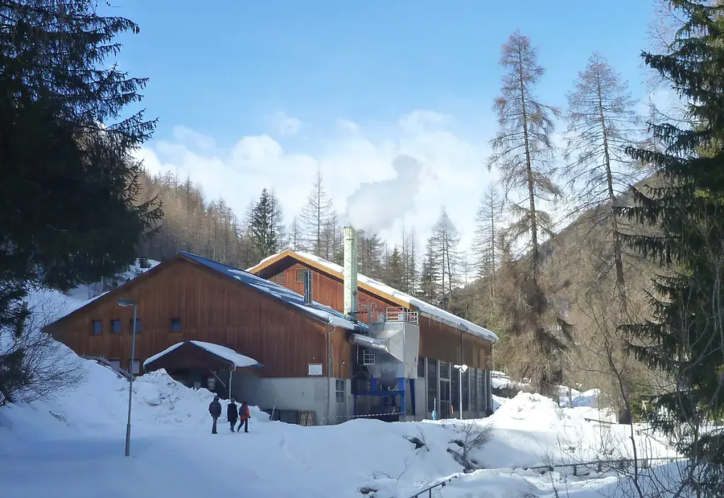 A timber-clad biomass heating plant with a chimney in a snowy Alpine valley